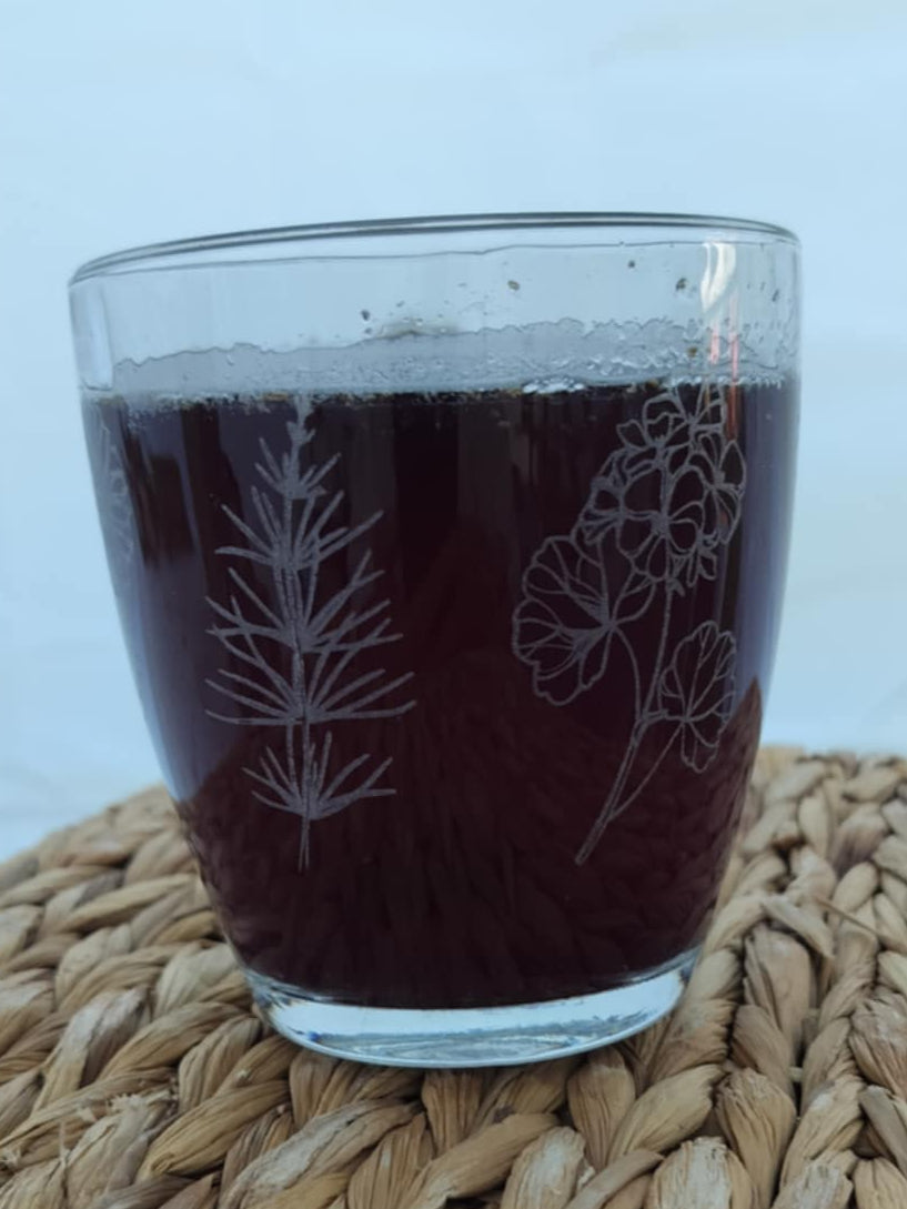 Glass of dark liquid with floral etching on a woven mat butterfly bliss herbal tea blend butterfly pea flower tea blue tea lemon verbena tea lavender tea tulsi tea holy basil tea nervous system tea postpartum tea nursing-friendly tea kid-friendly tea child-safe tea color-changing tea